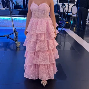 Rosa volangklänning i spets - Supersöt långklänning i ljusrosa spets med flera lager volanger och korsettband i ryggen. Klänningen är axelbandslös men finns möjlighet att lägga till band om man så önskar. Klänningen har en figurnära överdel med markerad byst. Perfekt för dig som vill sticka ut med en romantisk och drömmig look. Materialet är spets och modellen är riktigt prinsessig. ✨volangerna är uppsydda så att den passar längden för personerna i bilden. 💕