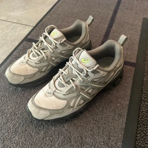  ASICS GEL NYC UTILITY skir strl 42,5 - Säljer ett par grå ASICS GEL sneakers med coola detaljer och svart sula. Skorna har snörning och meshpaneler för extra komfort. Perfekta för dig som gillar sportig stil och vill ha något som sticker ut med limegröna loggor.