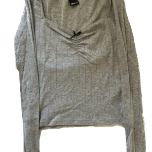 Grå ribbad croppad topp Gina Tricot - Grå långärmad croppad topp från Gina Tricot med smala vertikala ränder, en liten svart rosett framtill. Toppens material är mjukt och stretchigt, perfekt för en chill och snygg look.