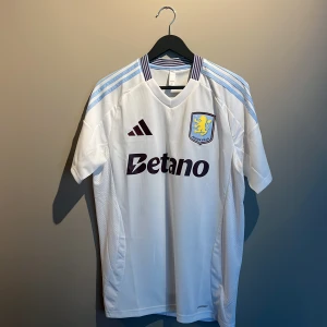 Aston Villa vit matchtröja Adidas M - Snygg Aston Villa fotbollströja från Adidas i vit med ljusblå och vinröda detaljer. Kortärmad med klubbmärke på bröstet och Betano-tryck. Tillverkad i lätt, ventilerande polyester med klassiska Adidas-ränder på axlarna. Perfekt för dig som vill representera Villa. Helt ny och oanvänd. Köpt på JD i London så 100% äkta!