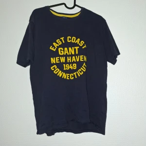 Mörkblå GANT t-shirt med tryck - Snygg mörkblå t-shirt från GANT med gul texttryck 'East Coast GANT New Haven 1949 Connecticut' på bröstet. Rund hals med gul detalj på insidan. Klassisk passform och korta ärmar, tillverkad i mjuk bomull. Perfekt för dig som gillar college-vibe och sportig stil.