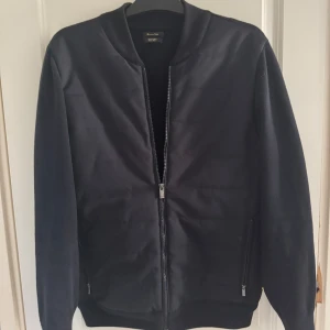 Massimo Dutti cardigan - Snygg svart bomberjacka från Massimo Dutti med stilren design. Jackan har dragkedja framtill, ribbade muddar och två fickor med dragkedja. Materialet är en mix av bomull och polyester som ger en mjuk och bekväm känsla. Perfekt för dig som gillar en clean look.