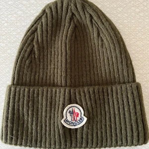 Olivgrön ribbad Moncler mössa - Snygg olivgrön mössa från Moncler med ribbad stickning och uppvikt kant. Märkets klassiska logga är broderad framtill. Perfekt för dig som vill ha en stilren och varm accessoar med exklusiv känsla.