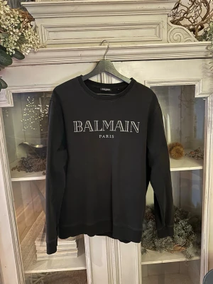 Svart sweatshirt från Balmain Paris - Svart sweatshirt från Balmain Paris med stor vit logotyp på bröstet. Tröjan har rund hals, ribbade muddar och är långärmad. Materialet är mjuk bomull som känns skönt mot huden. Perfekt för dig som gillar stilrena designer och vill ha ett statement-plagg.