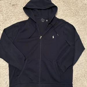 Svart hoodie från Polo Ralph Lauren - Svart hoodie från Polo Ralph Lauren med klassisk broderad logga på bröstet. Tröjan har huva med snörning, dragkedja framtill och två fickor. Tillverkad i mjuk bomull, perfekt för chill dagar. Snygg och enkel design som funkar till allt. Storlek L, har du funderingar är det bara att höra av sig! Priset är verkligen inte hugget i sten, pruta vid snabb affär!
