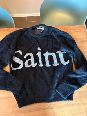 Svart stickad tröja Saintmex - Säljer en svart, supermjuk och fluffig stickad tröja från Saintmex Life Style. Tröjan har rund hals, långa ärmar och stor vit text 'Saint' framtill. Perfekt för dig som vill ha en statement piece med skön känsla.