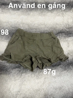 Olivgröna shorts med volangkant - Säljer ett par olivgröna shorts med elastisk midja och volangkant nertill. Shortsen har fickor på sidorna och en avslappnad passform, perfekta för varma dagar.🌸 Använd en gång. Storlek 98