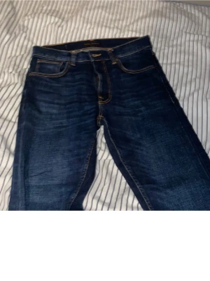 Mörkblå jeans från Nudie Jeans - Säljer ett par mörkblå jeans från Nudie Jeans med klassisk femficksdesign och snygga kontrastsömmar. Jeansen har en rak passform och är tillverkade i slitstarkt denim med Nudie-logga på bakfickan. Perfekta till vardags för en clean look.