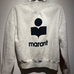 Grå sweatshirt från Marant, strl 36 - Säljer en ljusgrå sweatshirt från Marant i 100% bomull. Tröjan har ett stort svart Marant-tryck på ryggen och klassisk rund hals. Mjuk insida och ribbade muddar vid ärmslut och nederkant. Perfekt för en chill och stilren look.