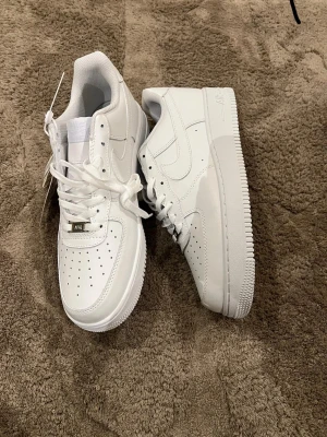 Nike Air Force 1 vita sneakers - Klassiska Nike Air Force 1 sneakers i helvitt skinn med perforerad tå, snörning och låg profil. Snygg och tidlös modell som passar till allt. Ikonisk Nike Swoosh på sidan och metallbricka med AF1 på snörningen.