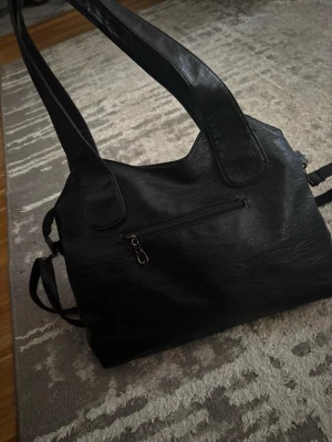 Svart axelväska i skinnimitation - Säljer en svart axelväska med breda axelband och flera dragkedjefack. Väskan har en stilren look och är gjord i skinnimitation med både korta och långa remmar. Perfekt för dig som vill ha plats för allt du behöver under dagen.