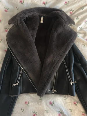 Svart teddyjacka från Mango - Säljer en svart teddyjacka från Mango med bred krage och mjukt, lurvigt foder. Jackan har flera silvriga dragkedjor och en croppad passform. Utsidan är i skinnimitation och insidan i mysig teddy. Perfekt för kalla dagar!