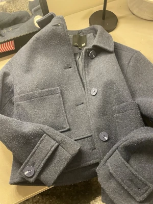 Blå ullrock med stora fickor - Snygg blå rock i ull med klassisk krage och stora framfickor. Jackan har knappar framtill och på ärmsluten, samt en stilren och rak passform. Perfekt för dig som gillar minimalistisk och tidlös stil. Kan sy på knappen !!