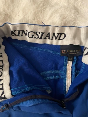Blå ridbyxor Kingsland strl 34 - Snygga blå ridbyxor från Kingsland med broderad logga på fickan bak och elastisk midja med Kingsland-tryck. Byxorna har förstärkta knäpartier och svarta detaljer vid bensluten. Perfekta för dig som gillar stilrena och funktionella ridkläder.