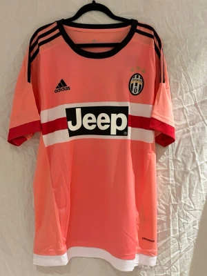 Juventus borta ställ 15/16 Pogba - Säljer en Juventus tröja i en snygg rosa färg med svarta och vita detaljer. Tröjan har svarta Adidas-ränder på axlarna, Juventus klubbmärke med tre stjärnor på bröstet och Jeep-logga framtill. Tillverkad i lätt och ventilerande polyester med climacool-teknologi. Perfekt för dig som hejar på Juventus och vill ha en unik och snygg tröja