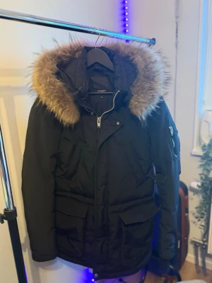 Svart parkajacka Rockandblue med päls - Säljer en svart parkajacka från Rockandblue med stor huva och fluffig päls. Jackan har dragkedja, knappar, flera fickor och en snygg detalj med dragkedja på ärmen. Perfekt för kalla dagar och riktigt skön passform.