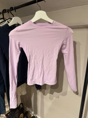 Ljuslila långärmad topp från Skims - En enkel och stilren långärmad topp i ljuslila färg. Modellen har rund halsringning och är figurnära utan några synliga detaljer eller mönster. Materialet känns mjukt och stretchigt, ganska genomskinligt. Perfekt att styla med jeans eller kjol. Från Skims.