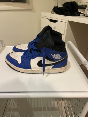 Nike Air Jordan 1 blå och vita sneakers - Säljer ett par Nike Air Jordan 1 sneakers i blått, vitt och svart. Skorna har klassisk hög siluett, blå snörning och svart swoosh på sidan. Ovandelen är i skinn och tungan är svart med blått Jumpman-logo. Perfekta för dig som gillar streetwear och ikoniska modeller.