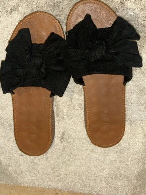 Svarta sandaler med rosett  - Säljer ett par sandaler med bred svart rosett upptill. Sulan är brun och har snäckmönster undertill. Öppen tå och platt sula gör dem perfekta för varma dagar. Materialet är syntet och textil, och designen är enkel men ändå söt.