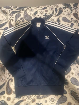 Mörkblå Adidas vindjacka S - Säljer en mörkblå Adidas vindjacka/ tjocktröja med klassiska vita ränder längs ärmarna och dragkedja framtill. Jackan har ståkrage, vita detaljer och Adidas-logga på bröstet. Perfekt för en sportig och clean look.