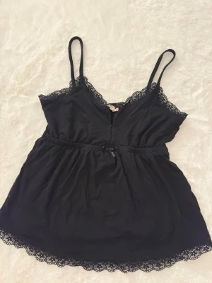 Svart spetsdetaljerad linnetopp - Säljer en svart linnetopp från Nelly i storlek XS. Toppen har tunna axelband, spetsdetaljer längs urringning och nederkant samt en liten rosett framtill i midjan. Materialet är mjukt och lätt, perfekt för varma dagar eller lager på lager.