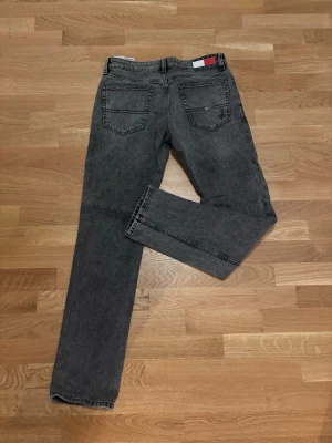 Tommy Jeans Slim grå jeans - Säljer ett par gråa Tommy Jeans Scanton Slim jeans med klassisk femficksdesign och diskret logga på bakfickan. Jeansen har en slim passform och raka ben, tillverkade i mjukt bomullsmaterial med snygg tvättad look.