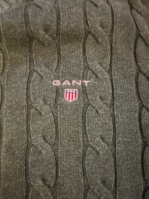 Grön kabelstickad tröja från GANT - Mörkgrön kabelstickad tröja från GANT i storlek XL. Klassisk rund halsringning och ribbade muddar vid ärmslut och nederkant. Broderad GANT-logga med sköld på bröstet. Perfekt för dig som gillar preppy stil och vill ha något både snyggt och bekvämt.