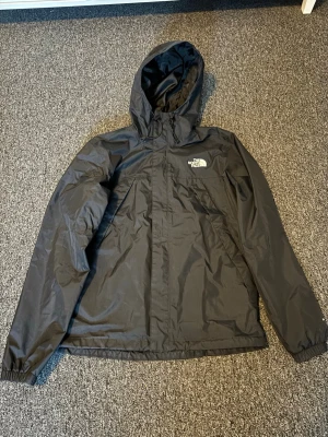 Svart vindjacka från The North Face - Svart vindjacka från The North Face med huva och dragkedja framtill. Jackan har en stilren design med logga på bröstet och meshfoder på insidan. 