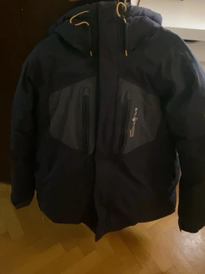 Svart pufferjacka från Sail Racing  - Säljer en svart pufferjacka från Sail Racing i storlek XL. Jackan har huva med gula snören, två vertikala bröstfickor med dragkedja och Sail Racing-logga på bröstet. Materialet är syntet och polyester, perfekt för kalla vinterdagar. Jackan är mindre i storlek. Jackan använder sig också av goretex system vilket gör att den också passar bra även om det regnar.