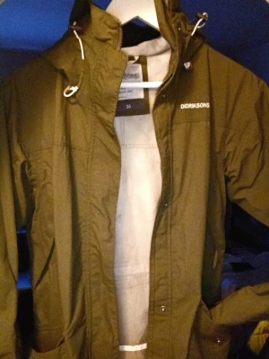 Grön vindjacka från Didriksons Thelma parkas - Säljer en grön vindjacka från Didriksons med huva och vita snören. Jackan har dragkedja framtill, praktiska fickor och en enkel, stilren design. Perfekt för blåsiga dagar och outdoor-äventyr. Passar dig som gillar funktionella plagg med snygg look.