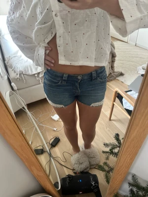 Blå jeansshorts med spetsdetalj - Säljer ett par blå denimshorts med låg midja och snygga spetsdetaljer vid bensluten. Klassisk femficksmodell med rå kant och dragkedja fram. Perfekta för sommaren och ger en avslappnad vibe.