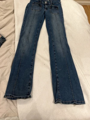 Blå bootcut jeans från Nelly - Säljer ett par blå bootcut jeans från Nelly med dubbla knappar i midjan och stora fickor fram. Jeansen har markerade sömmar längs benen och en klassisk tvättad look. Perfekta för dig som gillar retrovibbar och vill ha ett par jeans med lite vidare ben.