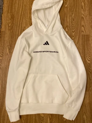  adidas hoddie  - Det är en vit hoodie från Adidas, gjord i ett mjukt och bekvämt tyg som ser både ren och välvårdad ut. På bröstet finns Adidas‑loggan placerad ovanför en vertikal text som lyder “ADIDAS SPORTSWEAR” i svart, vilket ger tröjan en sportig och modern känsla.