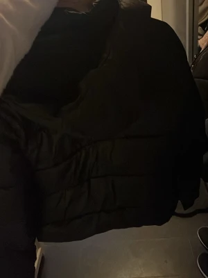 Svart pufferjacka med hög krage - Säljer en svart pufferjacka med hög krage och quiltad design. Jackan har en boxig passform och stängs med dragkedja framtill. Perfekt för kalla dagar när du vill hålla dig varm och samtidigt se cool ut.