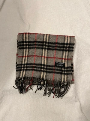 Burberry halsduk  - Klassisk rutig halsduk från Burberry i grått, svart, beige och rött. Tillverkad i 100% lammull och har fransar i ändarna. Perfekt accessoar för att lyfta din outfit med en tidlös brittisk vibe.