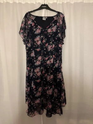 Blommig långklänning från Isolde - Supersöt långklänning från Isolde med mörkblå botten och blommigt mönster i rosa och vitt. Klänningen har v-ringning, volangdetaljer och korta, luftiga ärmar. Perfekt för sommarens alla tillfällen!