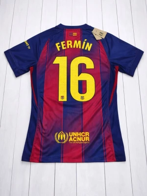 Barça matchtröja Nike Fermin #16 - Säljer en FC Barcelona matchtröja från Nike med Fermin och nummer 16 på ryggen. Tröjan har klassiska blå och röda ränder, gula detaljer och klubbmärke på bröstet. Tillverkad i lätt polyester med Dri-FIT ADV-teknologi och sponsorer som Spotify och UNHCR tryckta på framsidan.
