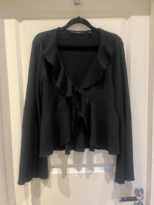 Svart omlottkofta från Vero Moda - Svart kofta från Vero Moda i omlottmodell med volangkant och knyt framtill. Långärmad och tillverkad i ett mjukt, ribbat material som ger en skön känsla. Perfekt att slänga över en topp för en chill och snygg look.