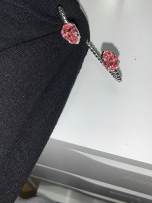 Silvrigt armband med rosa stenar från Caroline Svedbom - Säljer ett snyggt armband i tvinnad silverfärgad metall med två stora droppformade rosa stenar i ändarna. Armbandet är öppet och flexibelt, vilket gör det lätt att ta på och av. Perfekt accessoar för att lyfta din outfit.