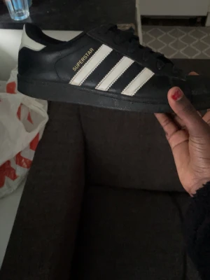 Adidas Superstar svart/vit sneaker - Klassiska Adidas Superstar sneakers i svart skinn med vita detaljer och de ikoniska tre ränderna på sidan. Skon har snörning, rund tå och vit häl med guldfärgad Adidas-logga. Perfekt för dig som vill ha en tidlös och streetinspirerad look.