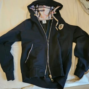 Burberry hoodie med dragkedja - Svart hoodie från Burberry med dragkedja och huva. Insidan av huvan har klassiskt rutigt foder i vitt, blått och rött. Tröjan har ribbade muddar, fickor framtill och ett broderat märke på bröstet. Perfekt för en chill och stilren look. Storlek M i tröjan men VÄLDIGT lite, jag skulle säga storlek xs. Köpte den här på plick i tron om att det var stl M. 