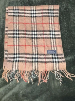 Rutig beige halsduk från Burberrys - Klassisk beige halsduk från Burberrys med ikoniskt rutigt mönster i svart, vitt och rött. Tillverkad i 100% lammull och har fransar i ändarna. Perfekt accessoar för att lyfta din outfit med en tidlös brittisk touch.