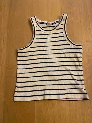 Randig ribbad linnetopp från Lindex - Säljer en vit ribbad linnetopp från Lindex med svarta horisontella ränder. Toppen har en klassisk rak passform och är ärmlös med breda axelband. Perfekt till sommaren och enkel att matcha med jeans eller shorts.