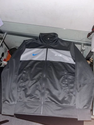 Nike Air svart och grå vindjacka - Nike Air vindjacka i svart och grått med blå swoosh på bröstet. Jackan har hög krage, hel dragkedja framtill och två fickor. Tillverkad i glansig polyester för sportig look. Perfekt för dig som gillar streetwear och sportig stil.