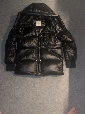 Svart Moncler pufferjacka med huva - Säljer en svart pufferjacka från Moncler med stor huva och blank finish. Jackan har Moncler-logga på bröstet, dragkedjor på fickorna och tryckknappar framtill. Perfekt för kalla dagar och riktigt snygg till streetwear-looken.