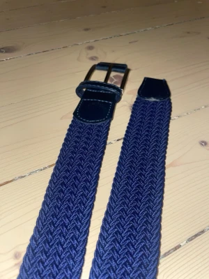 Mörkblå flätad bälte med skinn-detaljer - Snyggt mörkblått bälte med flätad design och detaljer i skinn vid spännet och ändarna. Enkel att justera och ger en stilren touch till outfiten.  105cm. Bara använt ett fåtal gånger.