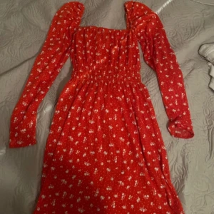 Röd blommig klänning  - Superfin röd klänning med små vita blommor och långa ärmad. Smockad överdel och markerad midja ger en snygg siluett. Perfekt för vår och sommar! Det blir kvadratringad vid halsen