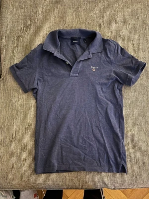 Marinblå pikétröja från Gant XS - Klassisk marinblå pikétröja från Gant i storlek XS. Tröjan har korta ärmar, krage och knäppning framtill. Gant-logga med broderad sköld på bröstet. Tillverkad i mjuk bomull som känns skön mot huden.