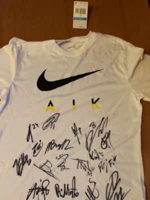 Signerad aik tröja från 2023 - Vit Nike Dri-FIT T-shirt med AIK tryck och signaturer. Storlek Medium.  Spelare som nummer 11 John Guidetti, nummer 47 Alexander fesshaie, nummer 45 Taha ayari, nummer 24 Omar faraj, nummer 20 zachary elbouzedi, nummer 43 Victor Andersson, nummer 35 Samuel Brolin, nummer 15 Kristoffer Nordfelt, nummer 5 Alexander Milosevic, nummer 4 sotiris papagiannopoulos, nummer 18 abdihakim ali och till sist nummer 23 budimir janosevic