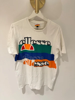 Vit t-shirt med tryck från Ellesse - Säljer en vit t-shirt från Ellesse med färgglatt tryck i grönt, blått och orange på framsidan. Klassisk rund hals och korta ärmar. Stort Ellesse-logotyptryck framtill ger en sportig vibe. Tillverkad i mjuk bomull för skön känsla.